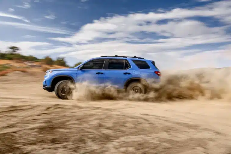 Blue SUV driving through sandy terrain with dust flying, under a partly cloudy sky.