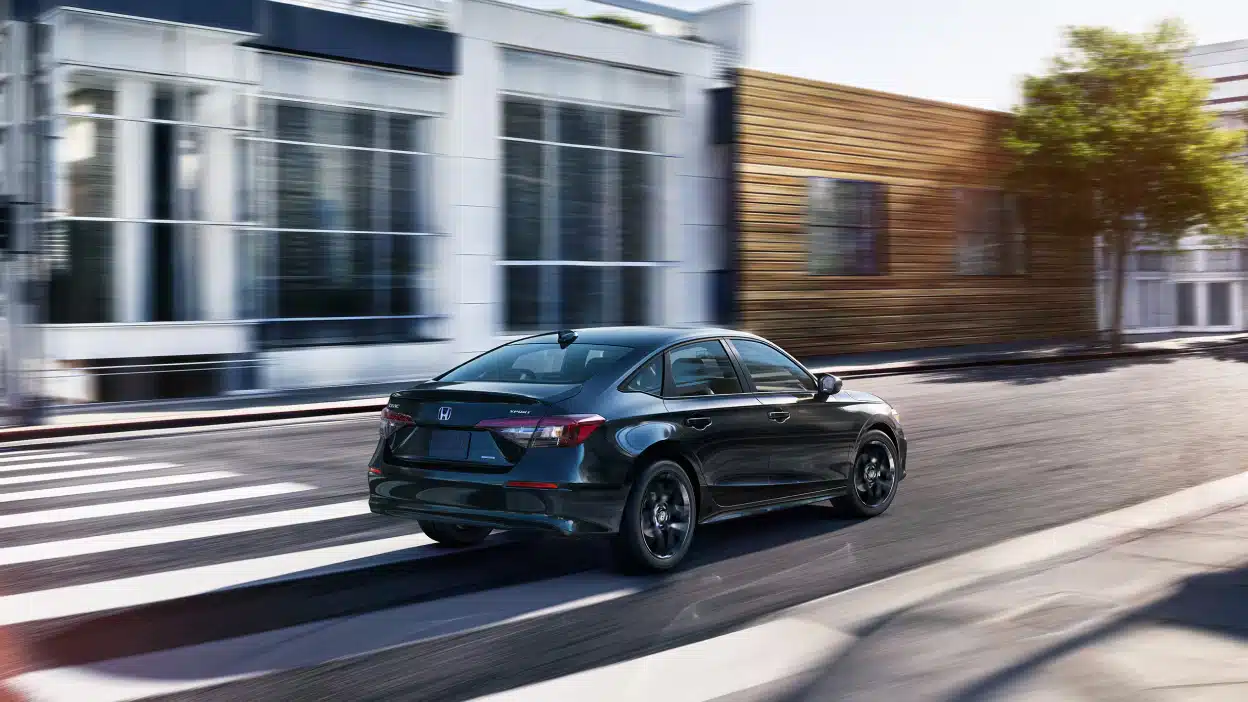 Black sedan driving on an urban road past modern buildings on a sunny day.