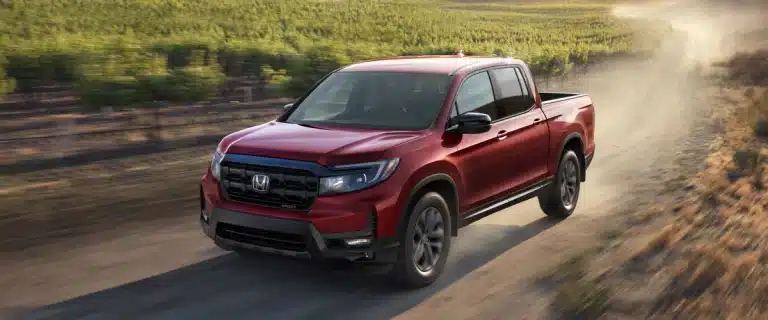 Red Honda pickup truck driving on a dirt road beside a vineyard under a clear sky.