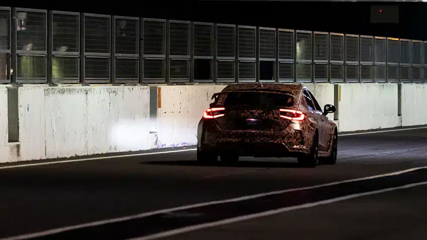 A camouflaged car with illuminated taillights on a dimly lit track, surrounded by barriers.