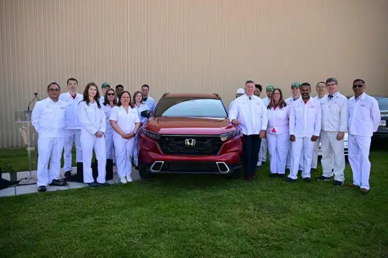 A group of people in white uniforms stand around a red Honda SUV, posing in front of a beige building.
