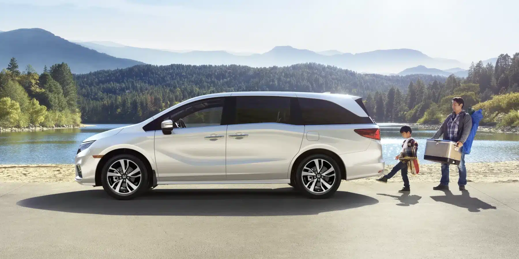 Silver minivan parked by a scenic lake with mountains, child and man carrying camping gear.