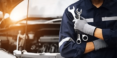 Mechanic in blue uniform holds wrenches, standing by an open car hood with engine visible.