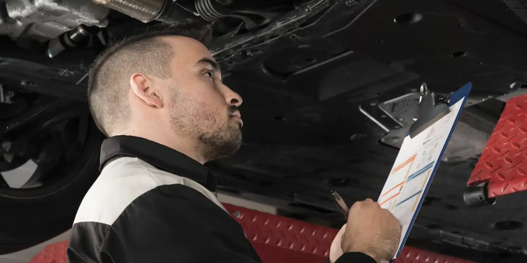 Mechanic inspecting a vehicle's undercarriage with a checklist on a clipboard for repairs.