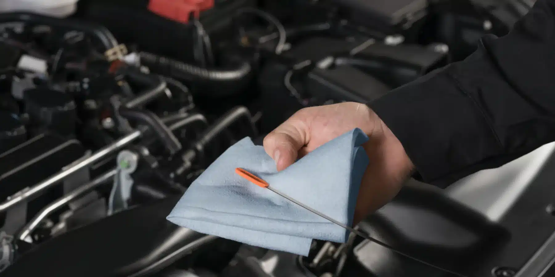 Hand holding a dipstick with blue cloth near an engine bay, checking vehicle oil.