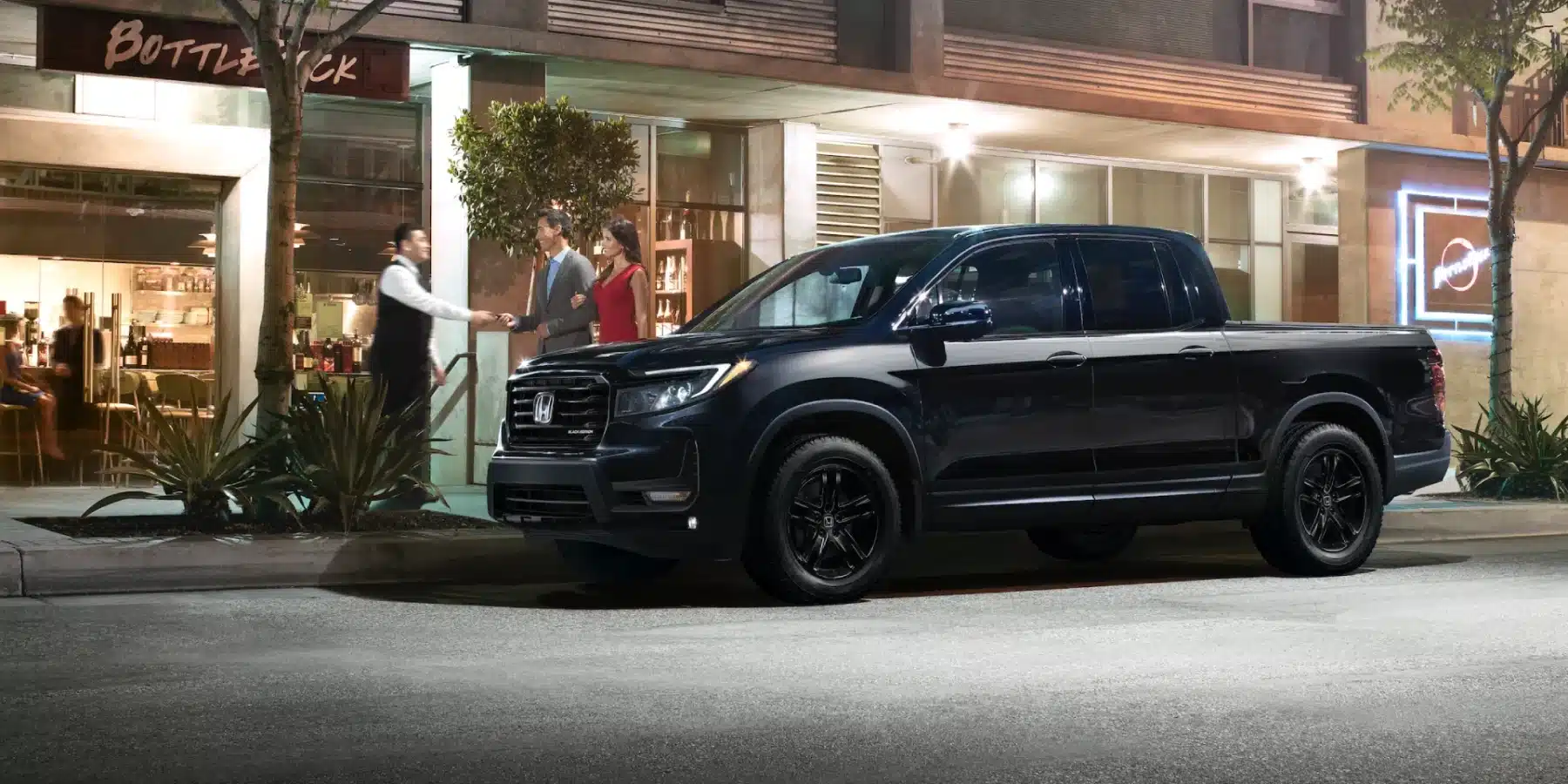 Black Honda Ridgeline parked outside a modern restaurant at night with people nearby.